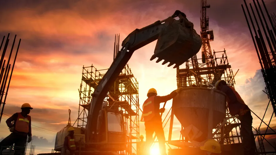 dramatischer Sonnenuntergangshimmel, im Vordergrund ein Bagger, im Hintergrund ein Kran auf einer Baustelle mit halbfertigen Gebäuden